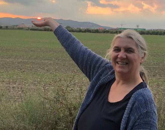 Doris Dvorak lächelt und zeigt mit ausgestrecktem Arm auf die Abendsonne über einem Feld